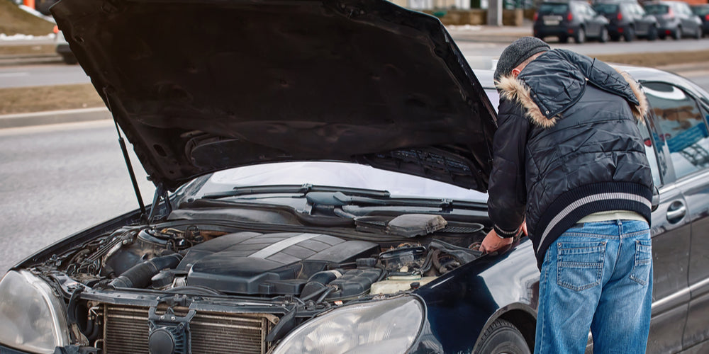 A sedan is parked in a grass bank on the side of a road with its hood open, while the driver stands in front inspecting the engine.