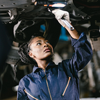 Female mechanic in blue coveralls working underneath a vehicle with a wrench.