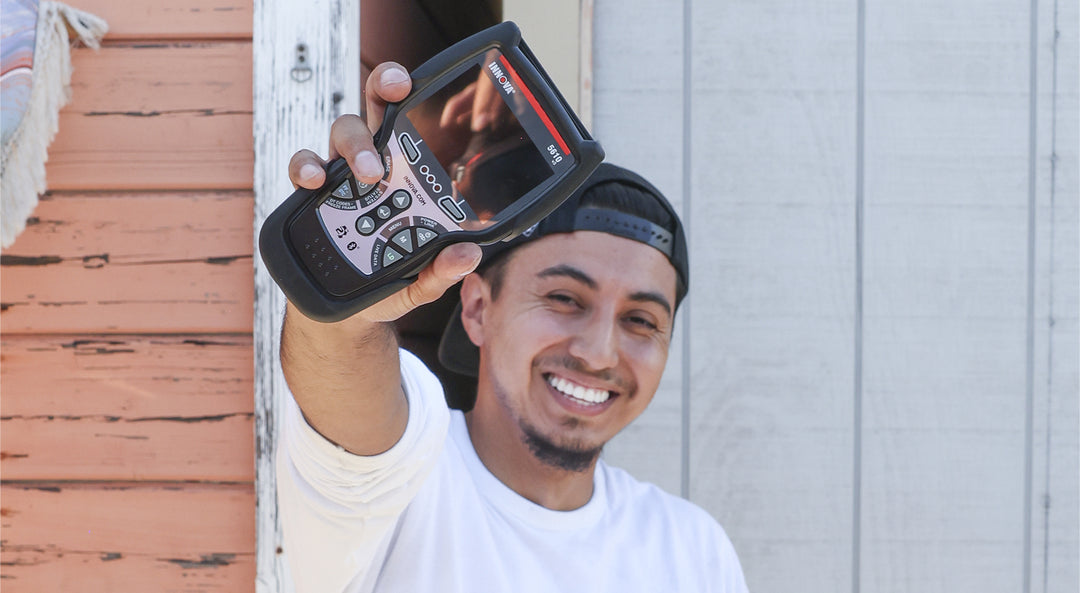 Smiling man in a white shirt and backward cap holding up a handheld device with display and buttons.